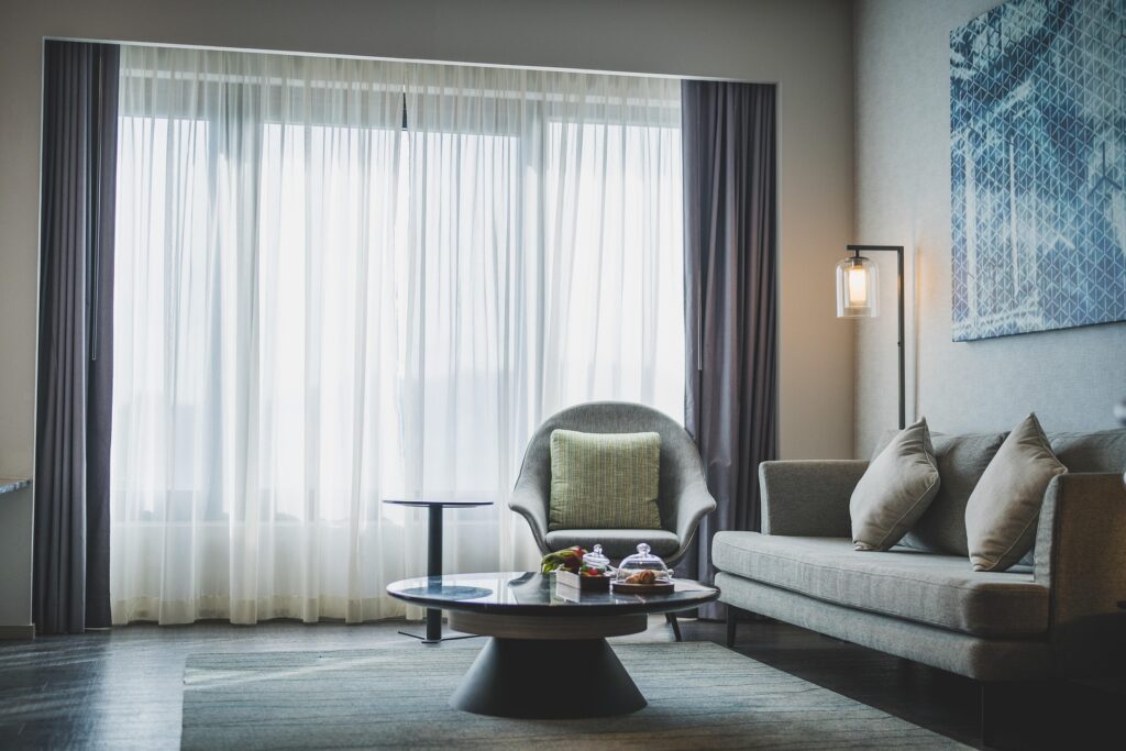 Zdjęcie przedstawia nowoczesny, elegancki salon z szarymi meblami wypoczynkowymi przy dużym oknie, w którym pod okrągłym stolikiem kawowym ułożono płasko tkany dywan w stonowanej kolorystyce.