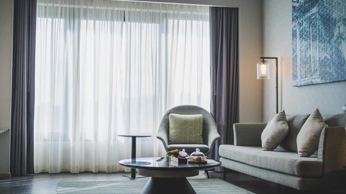 Zdjęcie przedstawia nowoczesny, elegancki salon z szarymi meblami wypoczynkowymi przy dużym oknie, w którym pod okrągłym stolikiem kawowym ułożono płasko tkany dywan w stonowanej kolorystyce.