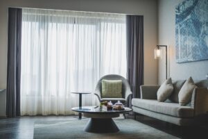 Zdjęcie przedstawia nowoczesny, elegancki salon z szarymi meblami wypoczynkowymi przy dużym oknie, w którym pod okrągłym stolikiem kawowym ułożono płasko tkany dywan w stonowanej kolorystyce.
